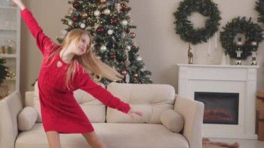 Kız Noel ağacının yanında neşeyle dans ediyor. Kırmızı kazaklı genç kız süslü bir Noel ağacı, şömine ve şenlikli çelenklerin yanında dans ediyor.