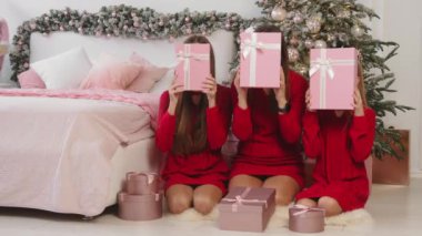 Kırmızı elbiseli üç kız Noel ağacının yanında pembe hediye kutuları taşıyorlar. Kırmızı elbiseli üç kız ellerinde pembe hediye kutuları, neşeli bir Noel ağacı ve rahat bir yatağın yanında oturuyorlar.