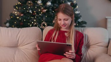 Noel ağacının yanında tablet kullanan kız. Kırmızı elbiseli genç kız arka planda güzel ışıklandırılmış bir Noel ağacı olan rahat bir koltukta tabletin tadını çıkarıyor.