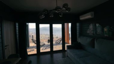 Dağ manzaralı samimi modern oturma odası. Cam kapılar ardında manzaralı bir dağ manzarası, şık bir dekor ve balkon olan karanlık bir minimalist oturma odasının içinde.