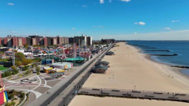 Sahil manzaralı, lunaparklı ve tahta kaldırımlı bir hava manzaralı canlı bir kumsal, hareketli bir eğlence parkı, bir sahil yürüyüşü ve açık mavi gökyüzü altındaki kıyı suları.