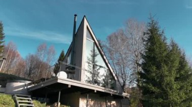 Cam Yüzlü Çerçeve. Ağaçlarla çevrili ve açık mavi gökyüzünün altında güneş ışığıyla yıkanmış büyük cam bir cepheye sahip modern A-frame kabin