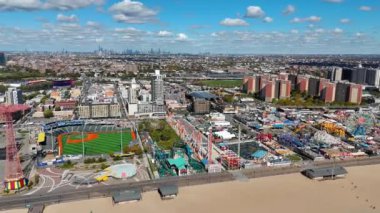 Lunapark ve şehir manzarasının havadan görüntüsü. Sahil kenarındaki bir lunapark, beyzbol stadyumu ve dağınık bulutlu mavi gökyüzünün altındaki şehir siluetinin geniş bir hava manzarası.