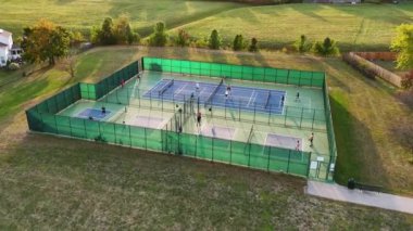 Açık Tenis ve Pickleball sahalarının hava görüntüsü. Yeşil alanlar ve ağaçlarla çevrili oyunların keyfini çıkaran oyuncularla çitle çevrili açık hava tenis ve yankesicilik sahaları.
