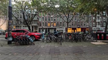 Hollanda, Amsterdam 29.11.2024 Timelapse Bisikletler ve Arabalar Şehir Caddesi 'nde park halindeki bisiklet ve park halindeki araçlar klasik tuğla binalar ve yemyeşil ağaçlarla çevrili..