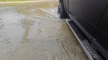 Sığ sularda giden kamyon, sığ sularda yüzen siyah bir kamyonun yakın görüntüsü engebeli bir off-road arazisinde dalgalanmalar ve sıçramalar yaratıyor..