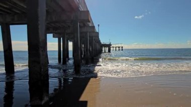 İskelenin altında, okyanus dalgalarının manzarası ve bir rıhtımın altından görünen kumlu sahil manzarası.