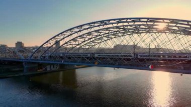 Krakow, Polonya 'da günbatımında nehrin üzerindeki kemerli çelik köprü. Gün batımında banliyö elektrikli treni bir köprüde gidiyor..