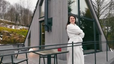 Beyaz cüppeli bir kadın ağaçlar ve karla kaplı tepelerle çevrili modern bir dağ kulübesinin güvertesinin keyfini çıkarıyor.