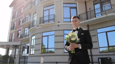 Damat elinde buketi, zarif modern binanın dış tarafında duruyor, siyah smokinli yakışıklı damat, modern bir binanın önünde beyaz bir çiçek buketi tutuyor..