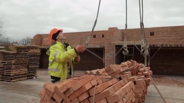 Tuğla kaldırma işlemini denetleyen inşaat işçisi, inşaat alanındaki tuğla paletini kaldırırken işçi rehberliği yapan vinç.