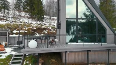 Ahşap bir terasta egzersiz yapan bir adam, büyük camlı pencereli modern bir A çerçeveli evin önünde. Teras, dağılmış ağaçları ve manzarası olan karlı bir yamaca bakıyor..