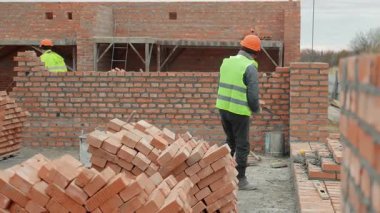 İnşaat işçileri tuğla duvar inşa ediyorlar. Güvenlik donanımlı iki inşaat işçisi inşaat sahasına tuğla duvar örüyor. Aletler ve tuğlalar görülebilir durumda.