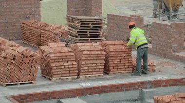 İnşaat alanında tuğla düzenleyen bir işçi. Güvenlik teçhizatı kullanan bir inşaat işçisi, geliştirme aşamasında olan aktif bir inşaat alanında ahşap paletlere tuğla yığını yerleştirir.