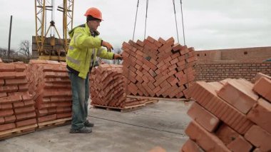 Vinç kaldıran bir işçi, bir palet tuğla kaldıran bir işçi, bir vinç tarafından kaldırılan tuğla paletine rehberlik eden bir inşaat işçisi.