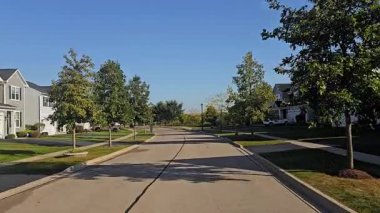 Sessiz bir banliyö sokağı, güneşli bir günde evler, ağaçlar ve açık mavi gökyüzü.