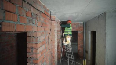 İnşaat halindeki bir binanın elektrik tesisatında çalışan bir elektrikçi güvenlik yeleği ve miğferli bir inşaat işçisinin tuğla bir duvara elektrik tesisatı kurması.