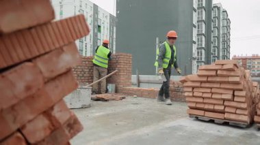 İnşaat işçileri dışarıda tuğla duvar inşa ediyorlar. Güvenlik donanımlı üç inşaat işçisi bir inşaat sahasına tuğla duvar örüyor. Aletler ve tuğlalar da yakınlarda..