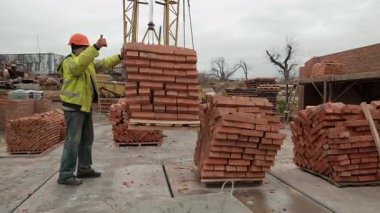 İnşaat işçisi vinç kaldırma tuğlaları, güvenlik ceketli bir inşaat işçisi ve vinçle tuğla kaldırmaya rehberlik eden bir inşaat işçisi.