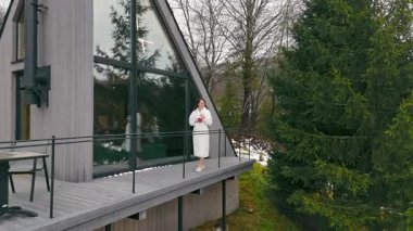 Beyaz cüppeli kadın modern kulübenin ahşap balkonunda elinde kırmızı bardakla doğa manzarasının tadını çıkarıyor.