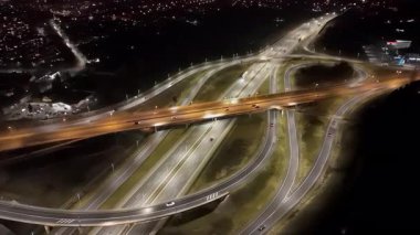 Gece aydınlık otoyol kavşağının havadan görünüşü, akan trafik ve çevre şehir ışıkları