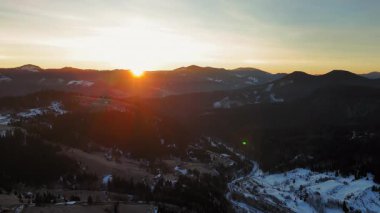 Karlı bir dağ vadisinde altın gün doğumu, sıcak bir sabah ışığı yuvarlanan tepelere ışık saçıyor, ormanları ve köyleri olan huzurlu kış manzarası..
