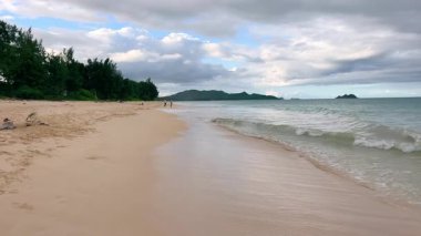 Sakin bir Hawaii plajı, yumuşak dalgaları, altın kumu nazikçe yıkayan dramatik bulutlar ve sakin okyanus ufkunu kaplayan uzak adalar.