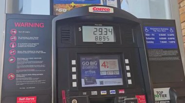 Chicago Illinois 05.03.2025 Costco benzin istasyonu benzin pompası arayüzü fiyat ve hacmi gösteriyor, uyarı işaretleri, ekran reklamları ve gündüz erişilebilirlik bilgileriyle çevrili.