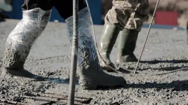 Kauçuk çizmeli işçilerin yakın çekimleri, döküm sırasında ıslak betonun içinde durmaları, elle inşa sürecinde güvenlik için bacaklara koruyucu bir örtü takmaları.