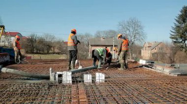 Turuncu yelekli inşaat işçileri bariyerlere beton döküyor, mavi gökyüzünün altındaki kırsal bölgedeki yerleşim alanında koordineli bir vakıf çalışması yapıyor.