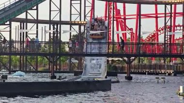 Polonya Energylandia 10.04.2025 Su yolculuğundaki insanlar lunaparktaki köprünün altına kırmızı lunaparktaki hız treniyle dalarken, Seyirciler yaz boyunca teknenin büyük bir dalga oluşturmasını izliyorlar.