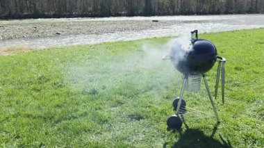 Açık hava barbeküsü sırasında çimenli nehir kenarında sigara içen yuvarlak kömür ızgarası, güneşli bahar havasında yükselen dumanla yemek pişirmek için taşınabilir çaydanlık ızgarası.