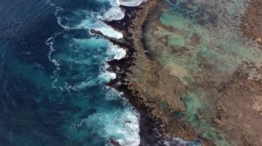 Mercan resiflerine çarpan dalgaların havadan görünüşü, turkuaz su ve kayalık kıyı görünür..