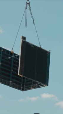 İnşaat vinci inşaat alanındaki büyük paneli kaldırıyor. Bir vinç, inşaat işçisi yüksek bir platformda dururken büyük bir paneli kaldırırken görülüyor..