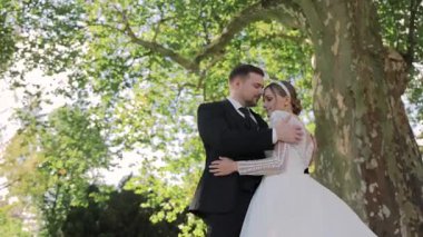 Gelin ve damat yaz ışığında büyük bir ağacın altında şefkatle kucaklaşıyorlar. Aşk dolu romantik bir an..