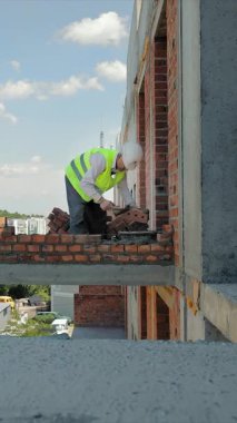 İnşaat işçisi, duvarlara tuğla yerleştiriyor. Yüksek görüş açısına sahip yelek ve sıkı bir şapka giyen bir inşaat işçisi tuğlaları binanın kenarına dikkatlice yerleştirir.