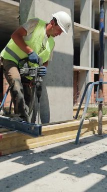 İnşaat işçisi inşaat sahasında sondaj yapıyor. Bir inşaat işçisi inşaat alanında yüksek görüş mesafesi olan bir yelek ve sıkı bir şapka takarak beton bir levhayı delerken görülüyor.