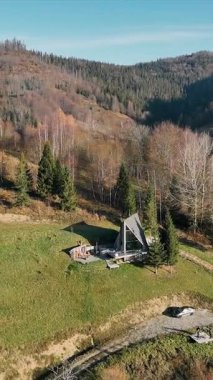 Sahne Manzaralı Hillside A-Frame Kabini. Çimenli bir yamaçta, ağaçlarla çevrili ve açık mavi gökyüzünün altında yuvarlanan tepelerle çevrili A çerçeveli bir kulübe.