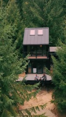 Ormanda, Noel ağaçlarının arasında, modern bir ahşap ev. İki adam terasta çay içiyor. Ormanda, inanılmaz doğanın ortasında rahat bir ev..