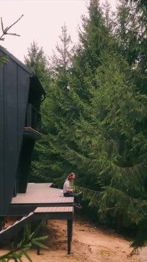 Yoğun Çam Ağaçları Arasında Yuvalanmış Kara Modern Kulübe. Parlak, siyah, modern bir kulübe balkon ile çevrili yoğun çam ormanına karışıyor, tenha ve sakin bir atmosfer yaratıyor güvertede sessizce oturan bir insan