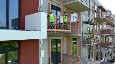 Askıya alınmış platformda inşaat işçileri cephe panelleri kuruyor. Etkin dış kaplama ve termal yalıtım aşamasının altındaki yeni apartman bloğu.