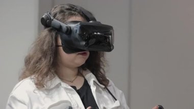 İçeride sanal gerçeklik kulaklığı kullanan bir kadın. Yüksek teknolojili VR kulaklık takan ve elinde kumanda olan genç bir kadın modern bir iç mekanda sanal bir deneyime daldı.