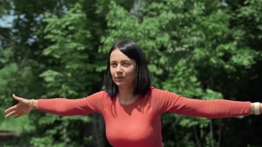 Dışarıda egzersiz yaparken kollarını açan bir kadın. Kırmızı bluzlu odaklanmış bir kadın, yemyeşil bir ortamda ısınma ya da yoga pozu verirken kollarını uzatır.