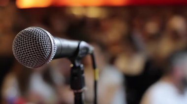 Arka planda bulanık seyirciyle Sahnede Mikrofonun Kapanışı, Topluluk Konuşmasını, Performans Gösterisini ya da Canlı Sunum Atmosferini simgeliyor.