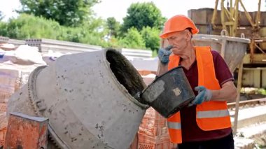 İnşaat Alanına Kask ve Güvenlik Yeleği Giyip, Beton Karıştırıcıya Su Döken Yaşlı İşçi.