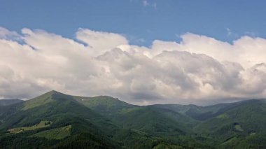 Bulutların zaman akışı yeşil dağ sırasının üzerinde kayıyor. Tüylü bulutların, yemyeşil dağ tepeleri ve orman vadilerinin üzerindeki mavi gökyüzünde hızla hareket ettiği hızlandırılmış görüntü.