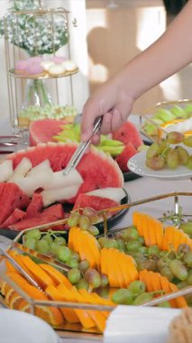 Bir adam ziyafet masasından meyve alıp tabağa koyar. Büfe masasında karpuz üzümü ve dilimlenmiş portakal.