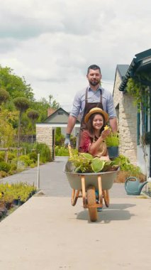 Mutlu bahçıvanlar, çiçeklerle dolu el arabasıyla bir kadını iten bir adam gibi birlikte gülerler. Neşeli enerjileri çiçek açan bahçeyi neşeyle dolduruyor..