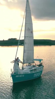 Drone gün batımında denizde yat yakalar. Gemi rengarenk bir gökyüzü altında sürüklenirken ışık sudan yansıyor..