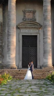 Yeni evliler, günbatımında çiçeklerle ve uzun antik sütunlarla çevrili eski bir kilisenin önünde sarılıyorlar. Aşkları akşam ışığında parlıyor.